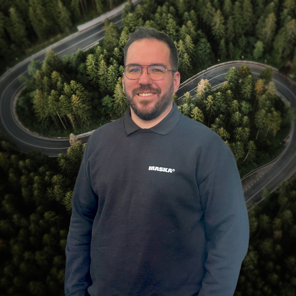 Homme portant un chandail noir MASKA, devant un décor aérien de route sinueuse traversant une forêt.