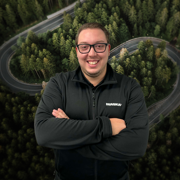 Homme portant un chandail noir MASKA, devant un décor aérien de route sinueuse traversant une forêt.