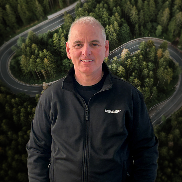 Homme portant un chandail noir MASKA, devant un décor aérien de route sinueuse traversant une forêt.